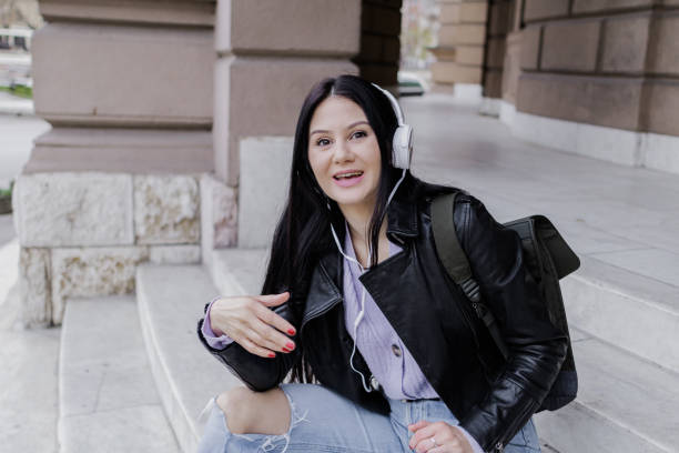 istock Young beautiful female university student with backpack sitting on college staircase. She is wearing headphones and leather jacket while waiting for her college class to start. 2091138731