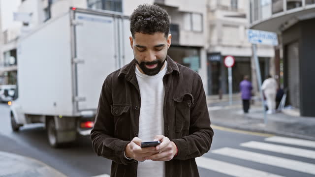 A young hispanic man with a beard using a smartphone on a bustling city street.