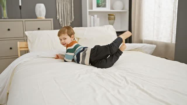 A playful boy in a striped shirt lies on a white bed in a cozy bedroom setting.
