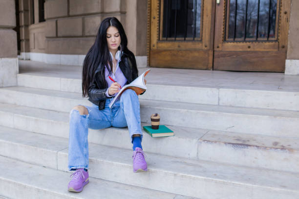 istock Young beautiful female university student with backpack sitting on college staircase and making notes with pencil, . She is wearing headphones and leather jacket while waiting for her college class to start. 2091150299