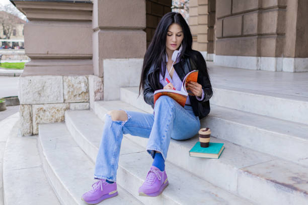 istock Young beautiful female university student with backpack sitting on college staircase and making notes with pencil, . She is wearing headphones and leather jacket while waiting for her college class to start. 2091150822