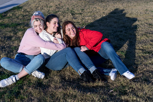 Three friends enjoy a relaxed moment sitting on the grass, basking in the sunlight, sharing smiles and a close bond in casual attire