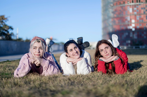 Three young women lying on their stomachs on the grass, posing playfully with hands under chins, with a clear sky and urban background