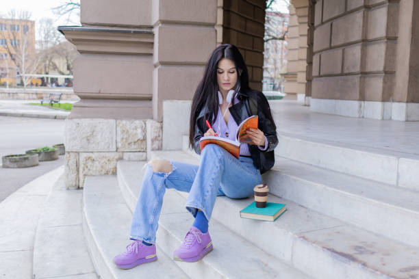 istock Young beautiful female university student with backpack sitting on college staircase and making notes with pencil, . She is wearing headphones and leather jacket while waiting for her college class to start. 2091154480