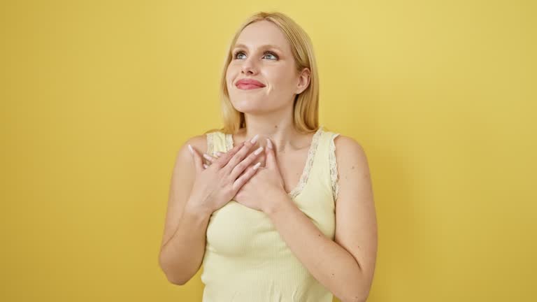 Beautiful young woman, blonde, sleeveless, standing over yellow background, wearing t-shirt, hands on chest, beaming with grateful health gesture. feeling proud, confident and cheerful.