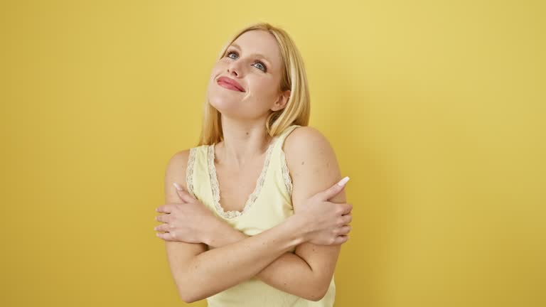 Beaming blonde beauty, young woman hugs herself in pleasure, wearing sleeveless t-shirt over yellow background. portrait of confidence, self love, and happiness.