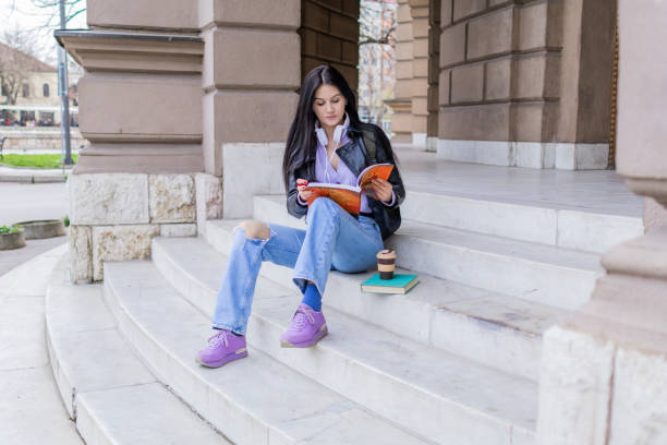 istock Young beautiful female university student with backpack sitting on college staircase and making notes with pencil, . She is wearing headphones and leather jacket while waiting for her college class to start. 2091159745