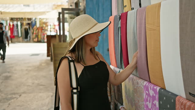 A woman in a wide-brimmed hat examines textiles at a colorful dubai market, evoking tourism and culture.