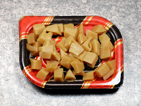 A Japanese delicacy, simmered bamboo shoots (筍), presented in a disposable plastic tray with a red and tartan border. The bamboo shoots are carefully cut into bite-sized pieces, highlighting their golden-yellow hue and tender texture. This dish is commonly found in delicatessens across Japan, offering a taste of seasonal produce and culinary simplicity.