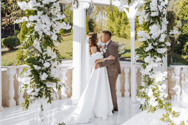 istock An elegant bride and groom pose together outdoors on a sunny wedding day against a background of flowers in a beautiful location. The groom gently hugs the bride and kisses her. 2091279451