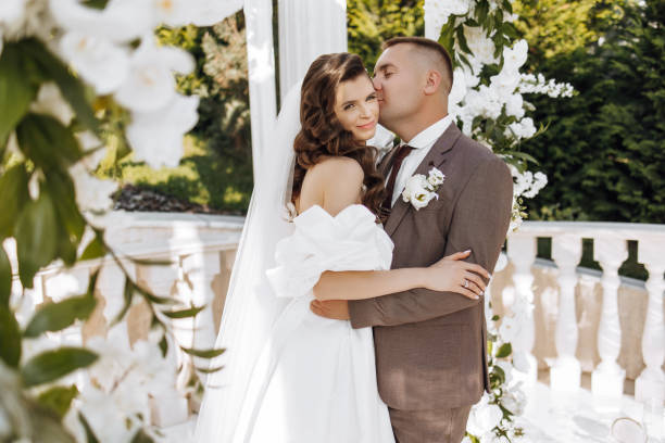 istock An elegant bride and groom pose together outdoors on a sunny wedding day against a background of flowers in a beautiful location. The groom gently hugs the bride and kisses her. 2091279455