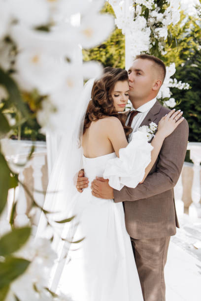 istock An elegant bride and groom pose together outdoors on a sunny wedding day against a background of flowers in a beautiful location. The groom gently hugs the bride and kisses her. 2091279459