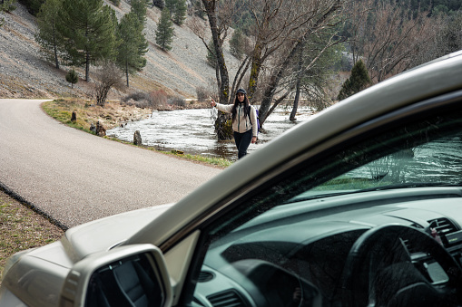 In the foreground of the image, a car is parked by a mountain road, while on the other side of the road, a woman hitchhiker is seen. The scene captures the encounter between two travelers amidst nature, with the car offering assistance to the hiker on her journey through the mountains