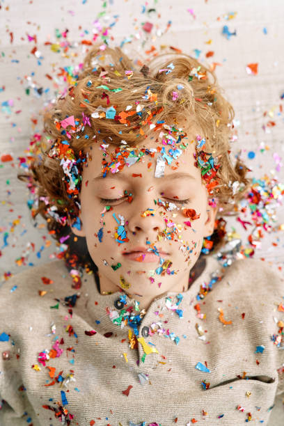 From above of adorable preteen boy lying on white surface with closed eyes and covered with confetti of poppers
