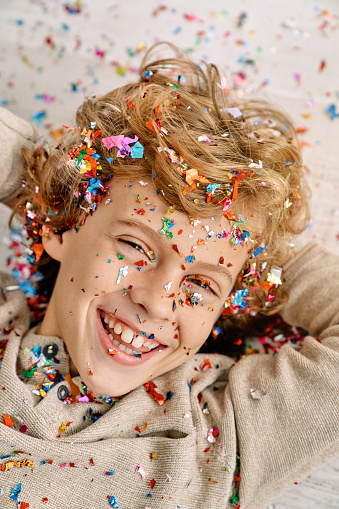 Happy blond kid smiling and looking at camera in confetti with hands on head