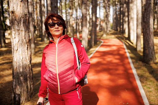 Middle aged woman carrying trekking poles while walking along red path in sunny forest within the tourist resort