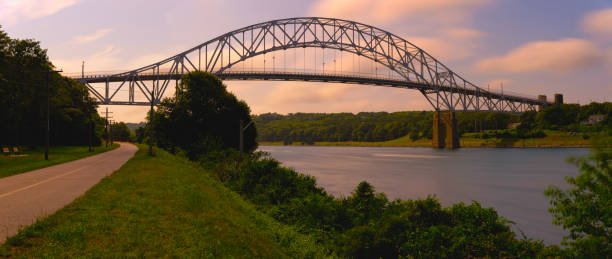 istock Cape Cod Canal Landscape with Sagamore Bridge at Sunrise with floating clouds and green bushes and trees along riverbank on Cape Cod, Massachusetts, USA, Long Exposure photo 2091418200