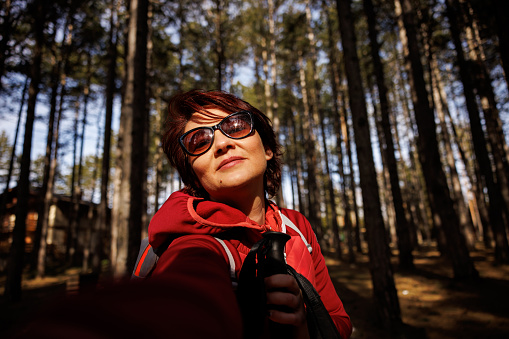 Smiling mature woman with sunglasses taking selfie in forest on sunny spring day, camera point of view