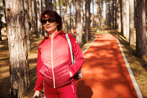 Middle aged woman carrying trekking poles while walking along red path in sunny forest within the tourist resort