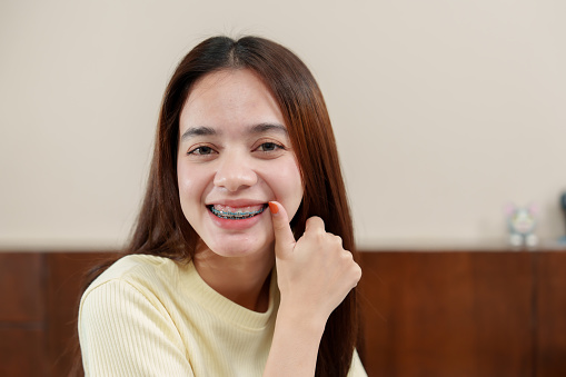 Asian female, pointing to braces, slight smile, cream top, clear focus on dental alignment, Eager adult showcases teeth straightening device, subtle grin, wooden furnishing completes homely scene