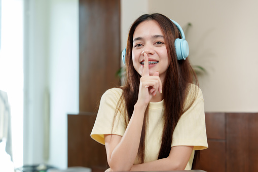 Asian lady with long hair, braces, wearing headset, gestures for silence, finger on lips, eyes gleaming with playful secrecy. Mirthful expression on face of person with dental braces, signalling hush