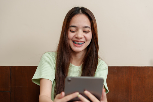 Engrossed young Asian lady with vibrant smile browses on tablet, casual attire adding to laid-back vibe. Concentrated female enjoys screen time, warm grin visible, relaxed ambiance in minimal setting.