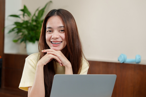 Young Asian woman with braces smiling at laptop, hands rested under chin, Joyful individual with dental orthotics engaged in remote work, propped elbows on table, houseplant and weights behind