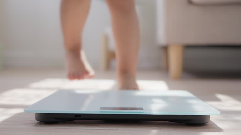 Little baby legs and barefoot walking step up standing on weight scales on wooden floor indoors at home. Children weight control concept.