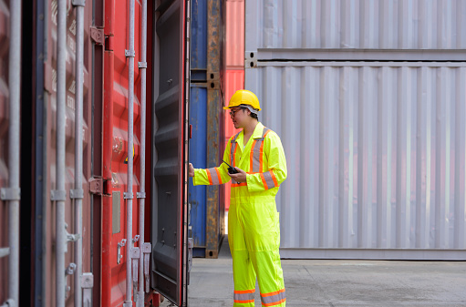 Portrait of Asian male worker wearing safety helmet and reflective vest opening container door for container inspection in freight cargo shipping yard