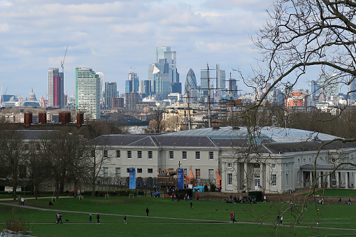 Greenwich, London, United Kingdom, March 16, 2024. View across Greenwich public park and historic buildings towards River Thames and distant Canary Wharf skyscraper. Spring day outdoors.