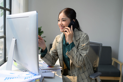 A woman is talking on her cell phone while sitting at a desk with a computer monitor in front of her