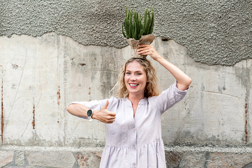 Happy blonde woman in pink dress holding pot of plant in craft paper on her head and showing OK sign gesture