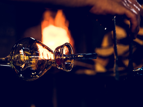 Pipe with shiny handmade glass bottle placed near burning flame of fire in dark glassblowing workshop