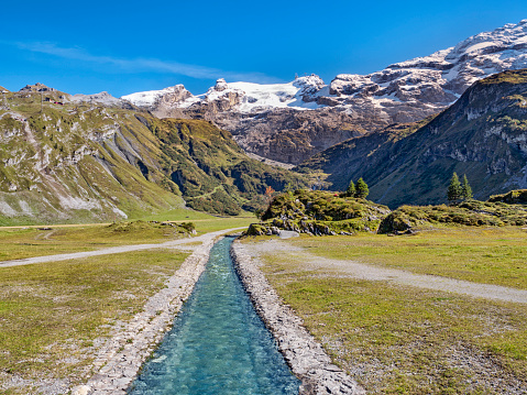 River stream in swiss alp mountains, beautiful green nature landscape, blue sky, sunny day, in the background snowy peaks of famous Titlis mountain, Switzerland