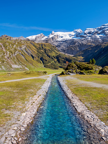 River stream in swiss alp mountains, beautiful green nature landscape, blue sky, sunny day, in the background snowy peaks of famous Titlis mountain, Switzerland