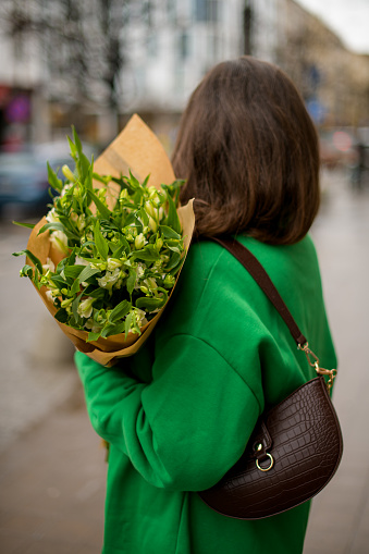 Turn away girl in a green jacket and a dark brown bag with a bouquet of incomparable flowers stands on the street