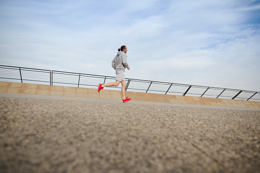 Side view of a determined muscular build young sportsman, male athlete jogger in gray active wear and red sports shoes, running on the city treadmill, enjoying morning jog on the promenade by bridge