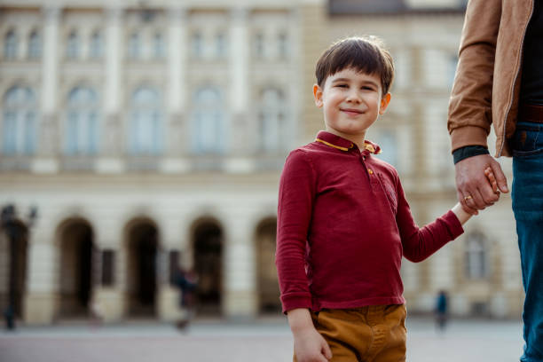 Cute Little Son Looking at Camera While Holding His Faceless Father Hand Outdoors in the City Center