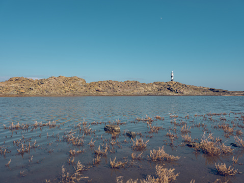 Scenic view of dry grass growing on sandy shore washed by blue calm sea and lighthouse on top of hill