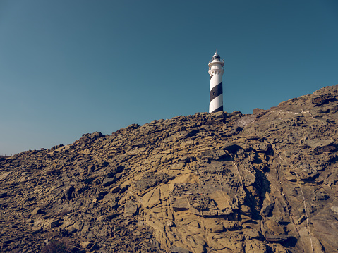From below of white lighthouse located on top of rough rocky formations under cloudless blue sky
