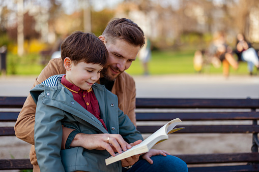 Portrait of Joyfull Young Father With His Adorable Little Son Reading Book While Sitting on the Park Bench. Family, Parenthood, Fatherhood, Adoption and People Concept