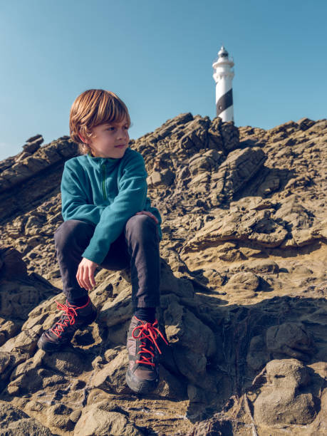 istock Boy sitting on rocks against lighthouse 2091752031