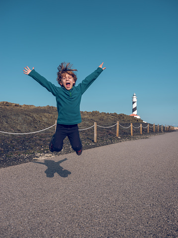 Happy little boy in casual clothes jumping with raised arms on dry walking leading to lighthouse under cloudless blue sky in daytime