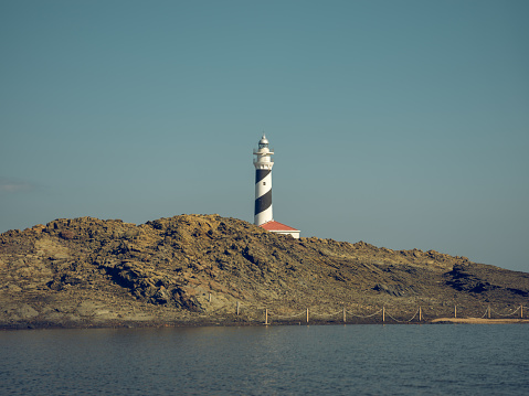 Lighthouse tower located behind rough mountainous coast of calm sea in sunny day