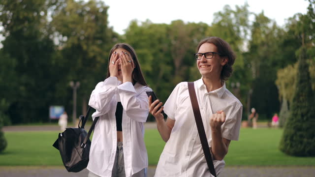 The guy shows the screen of his mobile phone to the girl and they look at it together, standing a city park. Having learned the results, the girl covers her face with hands, the guy jump and happy.