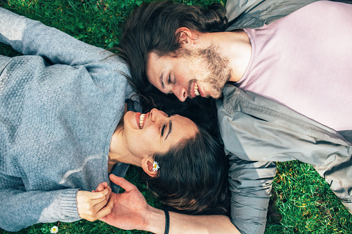 Smiling Young couple laying in grass, looking at each other and holding hands. They are beautiful looking happy and in love spending some time outdoor