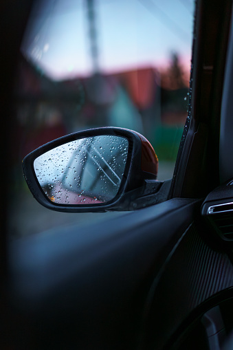 A car's rearview mirror viewed from inside the car while driving at dawn. The mirror is dotted with water droplets.