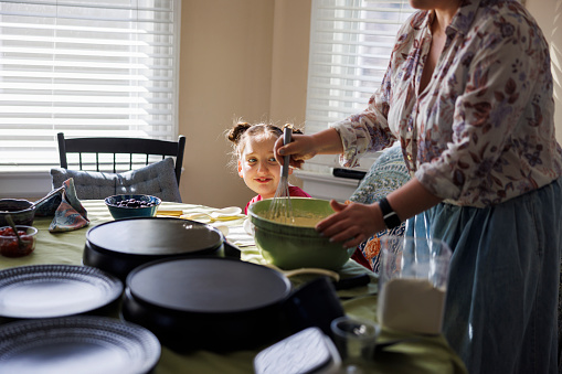 Cheerful young girl watching her mom preparing pancakes in a sunlit kitchen. Fried pans and plates in a kitchenware. Focus on a girl on background