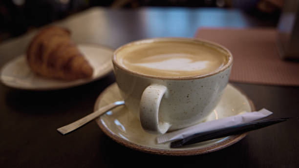 Delicious hot Italian cappuccino stands in a cup with a sugar pa Delicious hot Italian cappuccino stands in a cup with a sugar pa kets on a wooden table in cafe. In the background a fresh croissant on a plate spanish breakfast stock pictures, royalty-free photos & images
