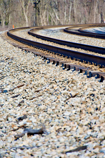 A set of curved railroad tracks slanted toward the inside of the curve atop a gravel railroad bed and railroad ties in a forested area on a sunny day.